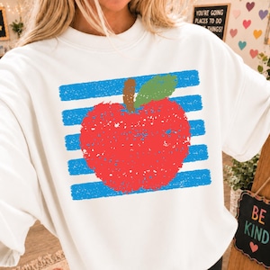 Peut inclure: Un sweat-shirt blanc à col rond arborant une impression graphique d'une pomme rouge avec une feuille verte centrée sur des rayures bleues horizontales. Le motif présente un aspect texturé et usé. La pomme est placée au milieu de la poitrine.