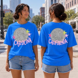 Può includere: Due donne indossano magliette blu brillante. La parte anteriore della maglietta dice "Carolina" in rosa, e la parte posteriore dice "California" in rosa. Ogni maglietta presenta una grafica a moneta con le parole "United States of America" e "Liberty".
