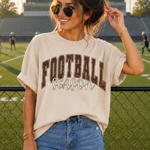 Może przedstawiać: Beżowy t-shirt z krótkim rękawem z napisem FOOTBALL SEASON, gdzie słowo FOOTBALL jest wykonane ciemnobrązową, postarzaną czcionką blokową, a słowo SEASON białą czcionką kursywną, zestawiony z niebieskimi dżinsami.