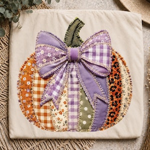 Op de afbeelding: Een beige t-shirt met een grote, centrale pompoenapplicatie gemaakt van diverse stoffen, waaronder oranje ruitjes, bloemenprints, luipaardprint en stippen. Bovenop de pompoen is een grote, paarse stoffen strik bevestigd.