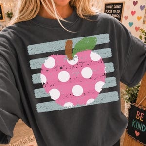 Peut inclure: Un sweat-shirt à col rond gris anthracite orné d'un grand motif de pomme rose vieilli à pois blancs. La pomme possède une tige brune et une feuille verte, superposée sur un fond de rayures horizontales bleu clair vieillies.