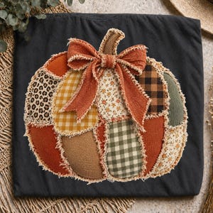 Può includere: Una t-shirt nera con una grande grafica centrale a forma di zucca realizzata con vari ritagli di tessuto dai bordi sfilacciati. Il design della zucca include motivi leopardati, quadretti gialli, fiori, scacchi neri e sezioni di tessuto arancione e verde.