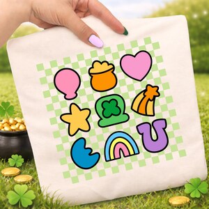 May include: A cream-colored tote bag featuring a green and white checkered pattern with colorful cartoon illustrations of a balloon, pot of gold, heart, star, shamrock, shooting star, horseshoe, rainbow, and crescent moon. The bag is held by a person.