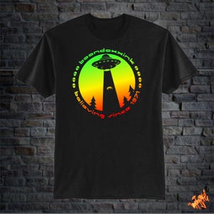 May include: Black t-shirt featuring a circular design with a UFO, trees, and the text "boondoxxink believing since 1971." The design has a gradient of green, yellow, and red. The UFO is above a beam of light.