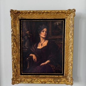 May include: A framed oil painting depicting a person in a dark velvet gown seated in a chair. The subject wears a dark necklace and is positioned against a dimly lit interior background. The artwork is encased in an ornate, textured gold frame.