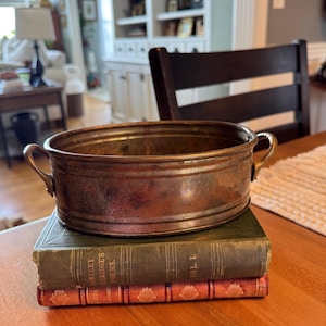 May include: A tarnished copper-colored oval bowl with two small handles, resting on a stack of two antique books. The books are bound in green and red with gold lettering. The bowl is approximately 12 inches long.