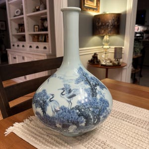 May include: A pale blue ceramic vase with a long, slender neck and a rounded body. The surface features a detailed blue underglaze illustration depicting multiple cranes standing among pine trees and rocky terrain, creating a scenic landscape.