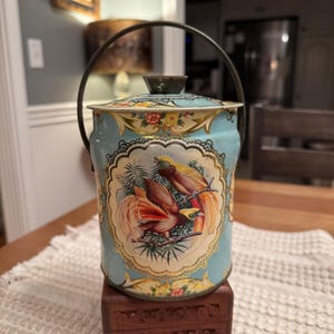 May include: Vintage metal cookie jar with a light blue background and a decorative bird illustration. The jar has a lid, a handle, and floral accents. The bird illustration features two birds with orange and yellow plumage.