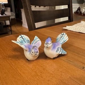 May include: Two ceramic blue jay figurines with outstretched wings and detailed feather patterns. The birds have white bellies, blue wings and crests, and black accents. They are displayed on a wooden table.