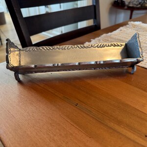 May include: A rectangular silver-toned metal tray with a smooth interior base and raised sides. The edges feature a repeating leaf-like embossed pattern. The tray is supported by four small, curved feet at each corner, elevating it slightly.