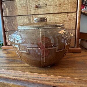 May include: A round, brown glazed ceramic casserole dish with a matching lid. The exterior features a raised geometric pattern consisting of intersecting lines and diamond shapes. It has two small side handles and a central knob on the lid.