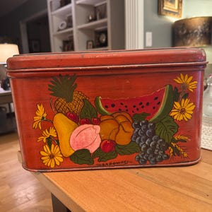 May include: A vintage rectangular metal tin with a hinged lid, painted red with a fruit design. The artwork includes a pineapple, watermelon, grapes, and other fruits and flowers. The tin is sitting on a wooden surface.