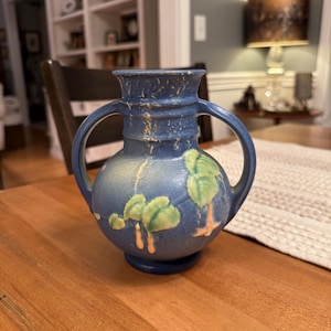 May include: A blue ceramic vase with two handles and a round body. The vase features green and orange tree designs. The vase is sitting on a wooden table.