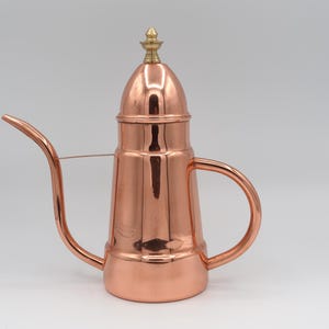 May include: A polished copper oil can featuring a tall, cylindrical body, a curved spout with a thin support wire, a rounded lid topped with a small brass finial, and a curved handle. The metal surface is reflective and smooth throughout.
