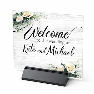 Pode incluir: Uma placa de casamento de acrílico transparente com as palavras "Welcome to the wedding of Kate and Michael" em escrita preta. A placa tem um fundo de grão de madeira branca e é decorada com detalhes florais. Está em um suporte preto.
