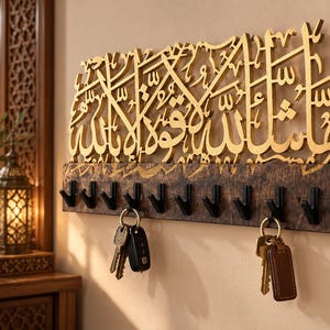 May include: A wall-mounted key holder featuring a dark, textured wooden base with ten black metal hooks. Above the hooks, a gold-colored cutout displays intricate Arabic calligraphy. Two sets of keys are hanging from the hooks on the rack.