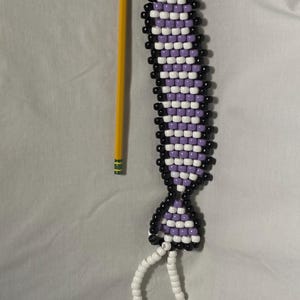 Puede incluir: Un marcapáginas de cuentas con un patrón morado, blanco y negro, que se asemeja a una corbata. Un lápiz amarillo está al lado del marcapáginas para comparar el tamaño. El marcapáginas tiene una cuerda de cuentas blancas.