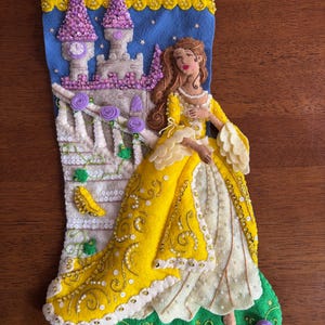 May include: A handmade Christmas stocking featuring a princess in a yellow gown, standing in front of a castle. The stocking is made of felt and decorated with sequins, beads, and embroidery. The background is blue with stars, and the bottom is green.