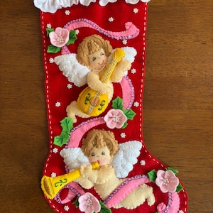 May include: Red felt Christmas stocking with embroidered angels playing musical instruments. The stocking is decorated with pink ribbons, flowers, and white trim. A festive holiday decoration.