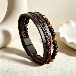 May include: A multi-layered bracelet featuring brown leather straps, a braided accent, and beaded strands. The bracelet incorporates black and tiger's eye beads, secured with a black magnetic clasp. The design is a blend of textures and colors.