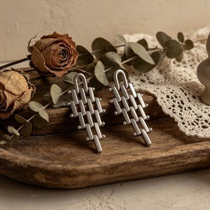 May include: A pair of silver-toned geometric drop earrings. The earrings feature a rectangular design with a series of connected bars and small round accents. The earrings are displayed on a wooden tray with dried flowers and greenery.