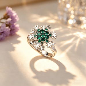 Könnte beinhalten: Ein silberner Ring mit einem Schneeflocken-Design. Das Zentrum des Rings ist mit einem Cluster aus kleinen, runden, smaragdgrünen Edelsteinen besetzt. Die Spitzen der Schneeflocke sind detailliert und filigran, was einen funkelnden Effekt erzeugt.