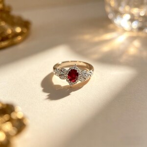 May include: A gold ring with a central oval-cut ruby gemstone. The ring features a detailed design with small, clear gemstones surrounding the ruby and along the band. The ring is set against a soft, neutral background, highlighting its elegant design.
