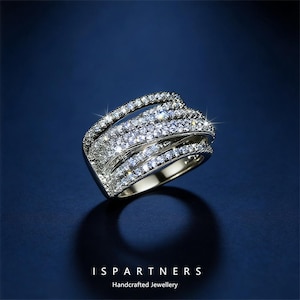 May include: A silver ring with multiple bands encrusted with clear, sparkling gemstones. The ring has a layered design, with the bands curving and overlapping. The ring is set against a dark blue background. The text "ISPARTNERS Handcrafted Jewellery" is at the bottom.