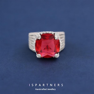 May include: A silver ring featuring a large, square, red gemstone. The ring has a textured band and small, clear accent stones around the red stone. The background is a dark blue. The text "ISPARTNERS Handcrafted Jewellery" is at the bottom.