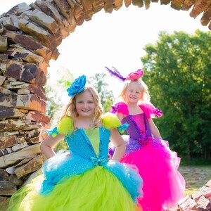 Pode incluir: Dois vestidos com saias de tule e corpetes. Um vestido &eacute; azul e verde-lim&atilde;o com um acess&oacute;rio de cabe&ccedil;a de flor azul. O outro vestido &eacute; roxo e rosa choque com um acess&oacute;rio de cabe&ccedil;a de flor roxa. Ambos t&ecirc;m saias de tule em camadas.