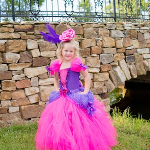 Pode incluir: Um vestido longo at&eacute; ao ch&atilde;o com uma saia de tule rosa choque e um corpete de croch&eacute; roxo com um painel central rosa brilhante. O vestido inclui mangas de tule bufantes, detalhes em fita roxa e um toucado a condizer com uma rosa rosa e uma pena roxa.