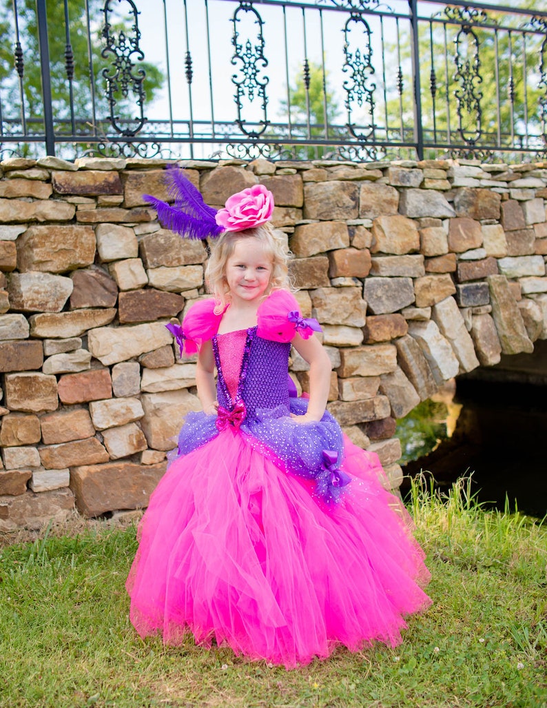 Pode incluir: Um vestido longo at&eacute; ao ch&atilde;o com uma saia de tule rosa choque e um corpete de croch&eacute; roxo com um painel central rosa brilhante. O vestido inclui mangas de tule bufantes, detalhes em fita roxa e um toucado a condizer com uma rosa rosa e uma pena roxa.
