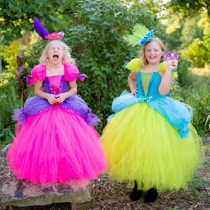 Pode incluir: Dois vestidos de tule at&eacute; ao ch&atilde;o. Um vestido apresenta um corpete roxo com uma saia rosa choque e um toucado de penas roxas. O outro vestido tem um corpete azul com uma saia verde-lima, um toucado de penas azuis e uma pequena coroa roxa.