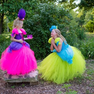 Pode incluir: Dois vestidos de tule. Um vestido apresenta um corpete roxo com uma saia rosa brilhante e um adorno de penas roxas. O segundo vestido tem um corpete azul com uma saia verde-lim&atilde;o e um adorno de flores azuis. Ambos os vestidos t&ecirc;m mangas bufantes.