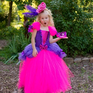 Pode incluir: Um vestido vibrante em f&uacute;csia e roxo com um corpete texturizado com detalhes em lantejoulas, mangas bufantes e uma saia de tule volumosa. O vestido &eacute; adornado com pequenos la&ccedil;os roxos e um acess&oacute;rio de cabe&ccedil;a de flor rosa com penas roxas.
