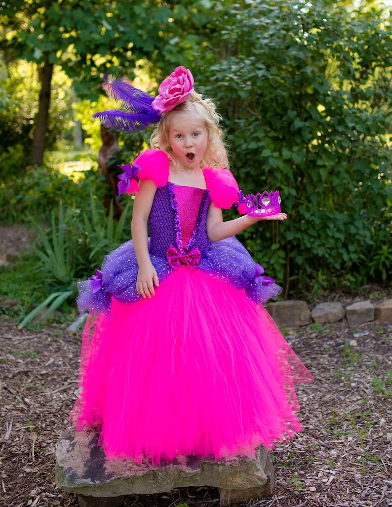 Pode incluir: Um vestido vibrante em f&uacute;csia e roxo com um corpete texturizado com detalhes em lantejoulas, mangas bufantes e uma saia de tule volumosa. O vestido &eacute; adornado com pequenos la&ccedil;os roxos e um acess&oacute;rio de cabe&ccedil;a de flor rosa com penas roxas.