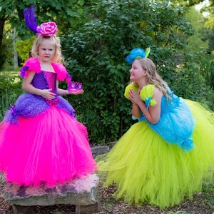 Pode incluir: Um vestido de tule roxo e rosa choque com corpete brilhante e um acess&oacute;rio de cabe&ccedil;a a condizer com uma grande flor rosa e uma pena. Um segundo vestido de tule azul brilhante e verde lima com um acess&oacute;rio floral azul a condizer.