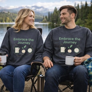 May include: Two people wearing dark gray sweatshirts with the text "Embrace the Journey" and a graphic of a notepad, pencil, plant, and clock. They are sitting in camping chairs near a lake, each holding a mug or tumbler.
