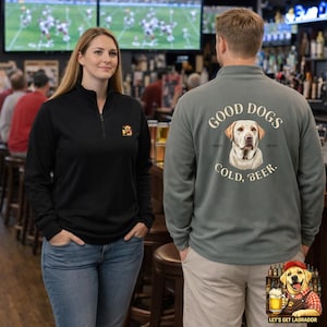 May include: Two people wearing black and gray quarter-zip pullovers. The black pullover has a small emblem on the chest. The gray pullover features a Labrador illustration with the text "GOOD DOGS, COLD, BEER."