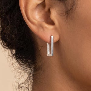 May include: A silver-toned rectangular hoop earring with a polished finish. The earring features a slim, elongated geometric shape that hangs vertically from the earlobe, showcasing a clean, metallic surface and a simple, hinged closure mechanism.
