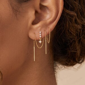Op de afbeelding: Een set goudkleurige oorbellen met fijne kettinkjes en staafvormige hangers. Eén oorbel bevat een verticale rij van drie kleine heldere steentjes en een stervormige bedel. De sieraden zijn ontworpen voor gebruik in meerdere gaatjes.