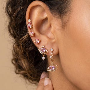 May include: A set of gold-toned earrings featuring pink heart-shaped stones. The designs include small heart studs, a heart surrounded by a ring, and a dangling heart charm with a star accent. Each piece is crafted with delicate metal details.