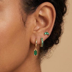 May include: A set of three gold-toned earrings featuring green stones. One earring is a small hoop with a dangling rectangular green stone, another is a small hoop with embedded green stones, and the third is a small stud with two marquise-cut green stones.