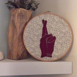May include: Embroidered hoop art with a burgundy hand making a wish with crossed fingers on a white background with black scribbles.