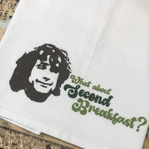 May include: A white tea towel with a black and white illustration of a man with long hair. The text "What about Second Breakfast?" is written in green and black lettering.