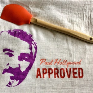 Puede incluir: Una toalla de cocina blanca con una silueta morada de la cara de un hombre y el texto "Paul Hollywood APPROVED" en rojo. Una espátula roja y de madera está encima de la toalla.