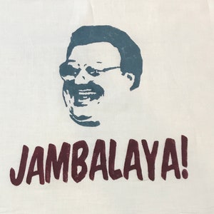 Könnte beinhalten: Ein weißes Küchentuch mit einer blauen Silhouette eines Mannes mit Brille und Schnurrbart. Der Text "JAMBALAYA!" ist in Rotbraun unter der Silhouette gedruckt.