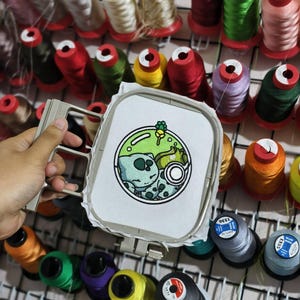 May include: An embroidery hoop holding white fabric with a circular design featuring a light blue and green creature resting inside a glass dome. The design includes a small green plant and a white circular icon. The hoop is set against a grid of thread.
