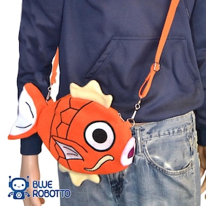 Puede incluir: Un bolso cruzado naranja con forma de pez y una larga correa naranja. El bolso tiene un ojo grande, aletas blancas y una aleta dorsal amarilla. El bolso se lleva sobre una sudadera con capucha azul y vaqueros. El texto "BLUE ROBOTTO" es visible.