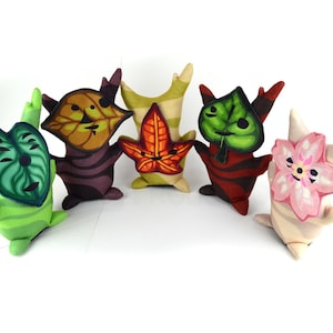 May include: A group of six plush toys, each shaped like a leaf or flower. The toys are in various colours, including green, orange, red, and pink, with detailed facial features. The plush toys are set against a white backdrop.